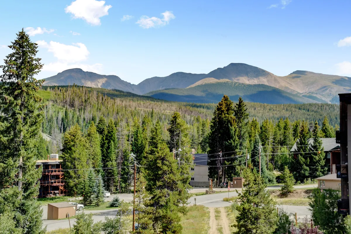 A panoramic view of a mountain range at sunset with a dense forest and a winding road leading into the distance. Buildings sit in the foreground, inviting visitors to explore nearby trails, go hiking, and enjoy peaceful moments in nature.