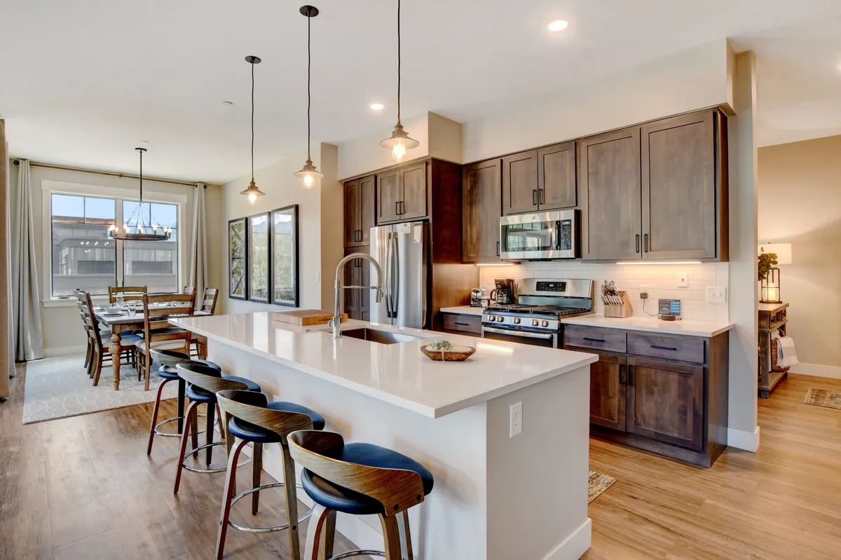 This interior image centers on a spacious, modern kitchen with a sizable island at its heart. The kitchen is accented by sleek cabinetry, a prominent cooking area, and stools pulled up to the island, all set against the backdrop of a large, sunlit window. The dining table is nearby, completing the sense of an open, communal space designed for gatherings, cooking, and entertaining.