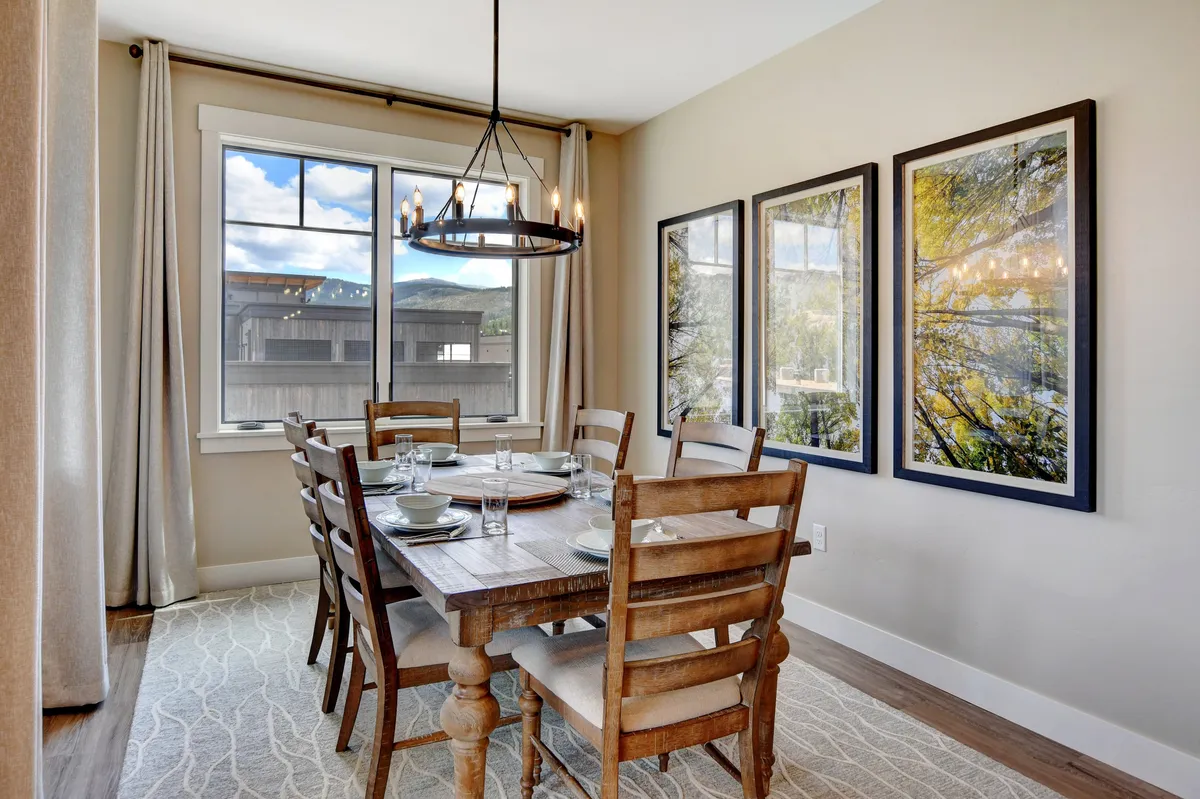 This image highlights a cozy dining area with a round table set beneath a statement wagon wheel chandelier. Framed artwork and a large window add character and brightness to the space, making it ideal for family gatherings and festive occasions.