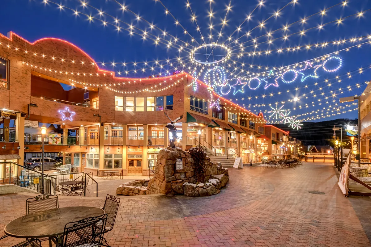 The image features an outdoor seating area illuminated by string lights, decorative stars, and a stone fountain, creating a magical and joyful ambiance. People are seen enjoying the space, which is ideal for social gatherings, dining, or celebrating community events.