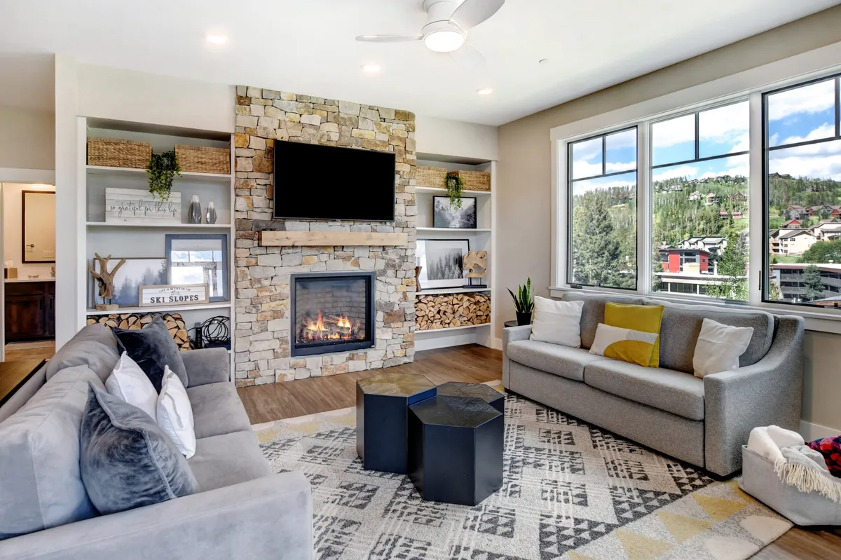 The image showcases a stylish, open-concept living area featuring a striking stone fireplace as the centerpiece, surrounded by plush, comfortable seating. Expansive windows flood the space with natural light and offer sweeping views, while decorative shelves add a touch of mountain lodge charm. The room radiates warmth and relaxation, inviting guests to gather, unwind, and soak in the cozy ambiance.