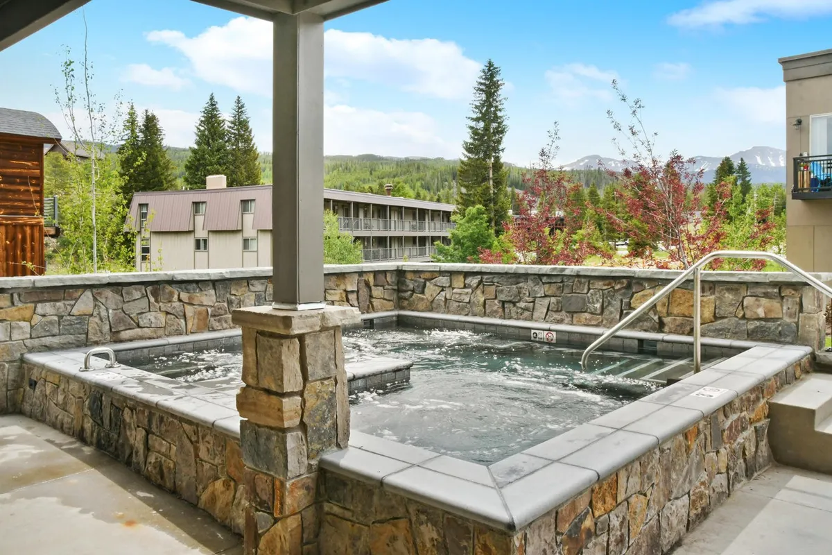 An exterior community hot tub surrounded by a stone wall, set against a backdrop of trees and mountain scenery. The inviting water, outdoor ambiance, and natural setting create a relaxing, resort-like atmosphere.