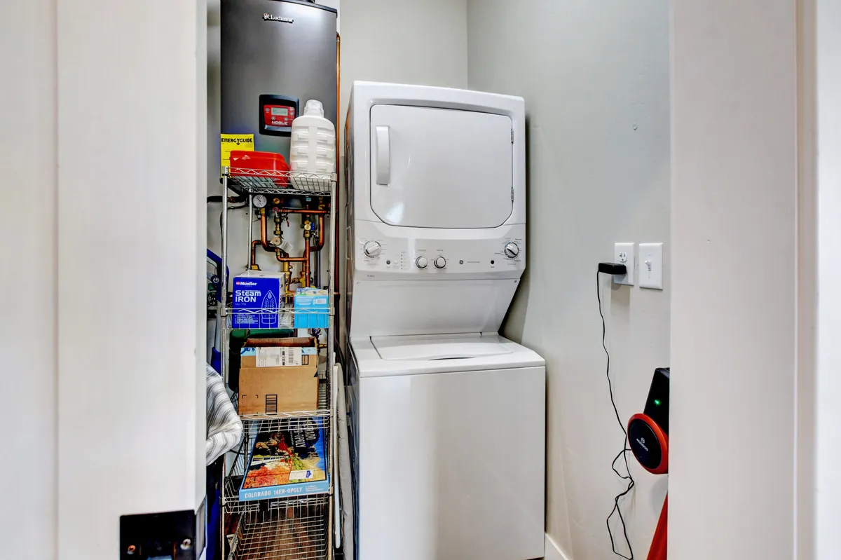 A compact in-unit laundry area featuring a stacked washer and dryer, storage rack, and household items organized for easy access. The space is clean and highly functional, ideal for longer stays and families.