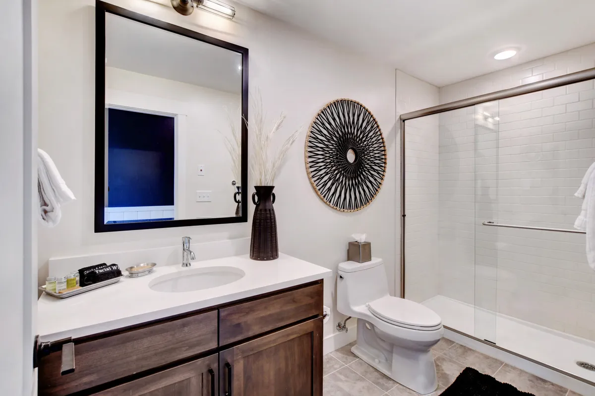 A modern, serene bathroom featuring a large mirror, decorative wall art, a spacious shower enclosure, a vanity sink, and a toilet. Soft lighting enhances the spa-like feel, perfect for self-care routines.