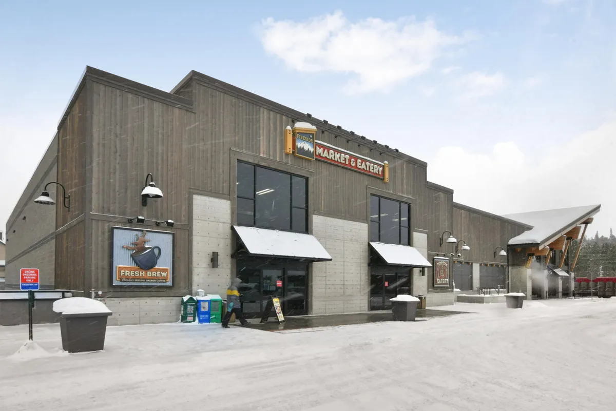This image features the exterior of a local market with a welcoming sign, entrance doors, and a snow-covered ground. People are engaged in grocery shopping, casual dining, and socializing with friends outside, all set in a cozy, wintery atmosphere. The setting hints at local community life and the convenience of nearby amenities for social gatherings.