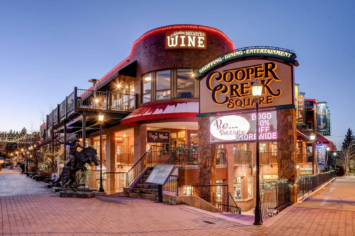 A lively street scene at Cooper Creek Square, featuring a well-lit sign, upper deck with lights, various storefronts, and a welcoming statue at the entrance. The area is animated with visitors, suggesting shopping, dining, and entertainment opportunities.