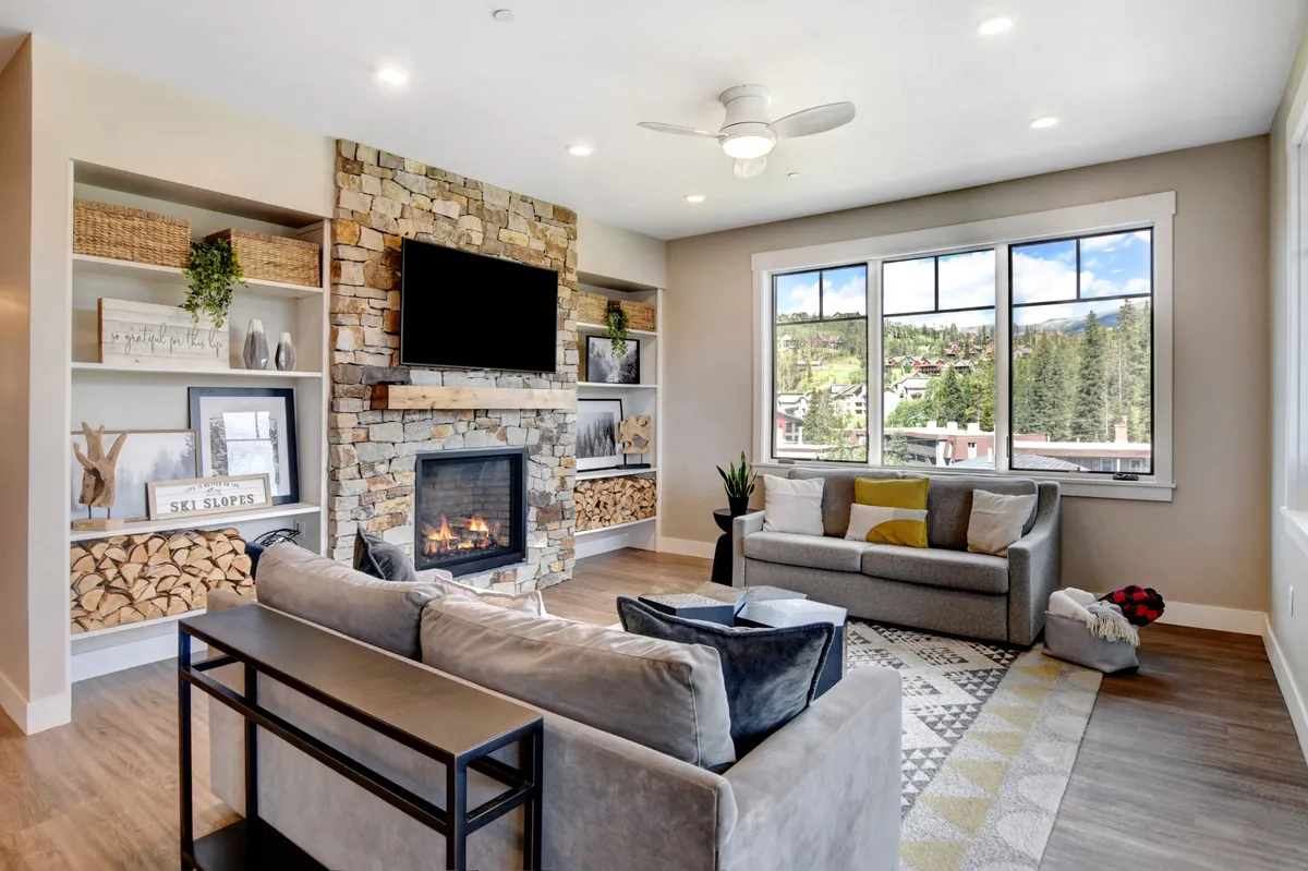 This inviting interior image features a large fireplace, comfortable seating, decorative shelving with games, and expansive windows looking out onto mountain views. The warm, welcoming space is perfect for gathering, socializing, and relaxing after a day of exploration.