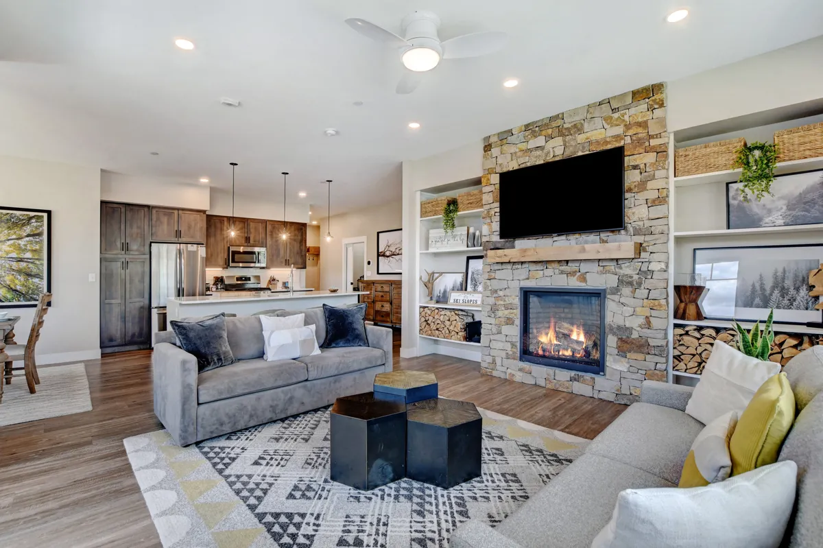 A welcoming open-concept living area featuring a stone fireplace as the focal point, flanked by comfortable sofas, a vibrant rug, and a kitchen visible in the background. The arrangement encourages social interaction, relaxation, and entertainment, with a cozy, lived-in atmosphere ideal for family gatherings.