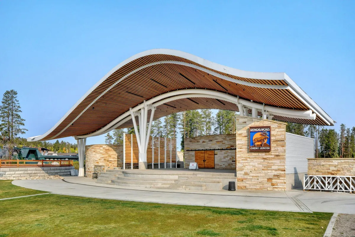 The exterior showcases a striking curved roof structure, a stone facade, and an open stage area designed for outdoor events, concerts, and community gatherings. The festive mood and ample space reflect opportunities for group engagement and memorable experiences under the stars.